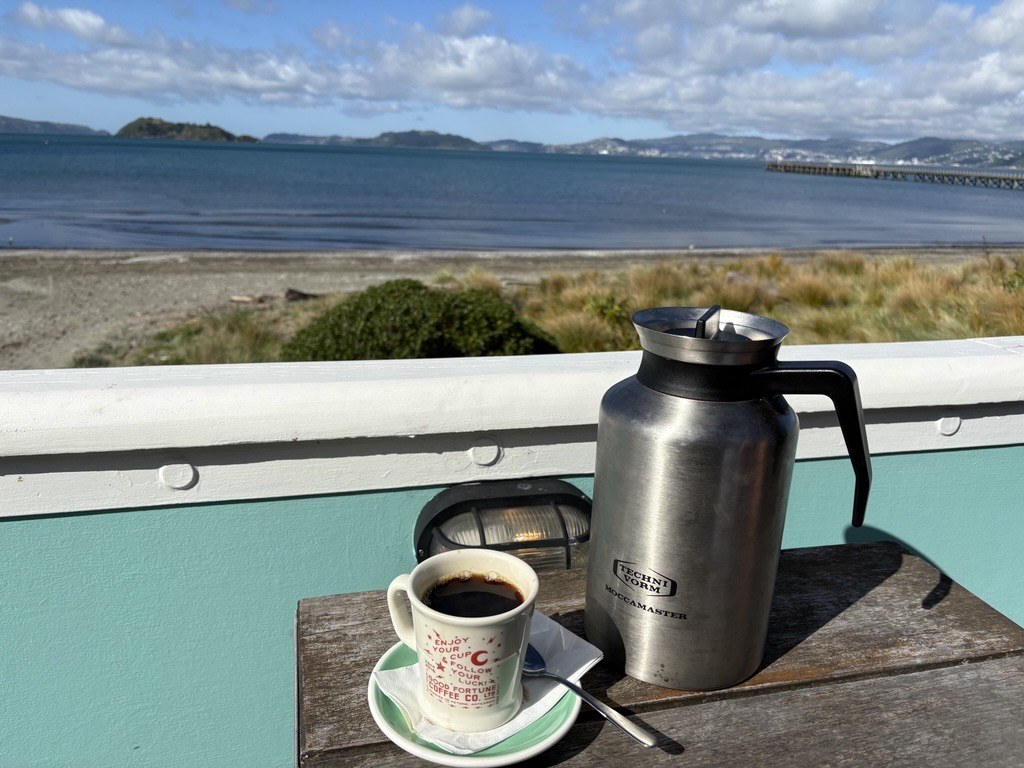 A cup of coffee and coffee pot on a table overlooking the beach and water